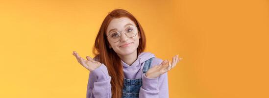 Nothing much. Attractive silly unsure carefree redhead unbothered cute female wearing geek glasses smirking uncertain shrugging hands spread sidways clueless what do future, orange background photo