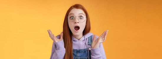 Close-up shocked sensitive concerned young panicking redhead woman worry drop jaw gasping raise hands spread freak out stare surprised emotional reacting incredible news, orange background photo