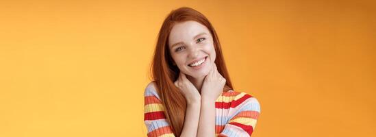 Silly enthusiastic attractive redhead blue-eyed girl tilting head touching neck flirty smiling enjoy perfect day feel happiness joy giggling coquettish flirting boyfriend, standing orange background photo