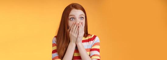 Shocked upset redhead female witness terrible accident gasping cover mouth palms astonished staring left frightened express empathy worry standing orange background panicking photo