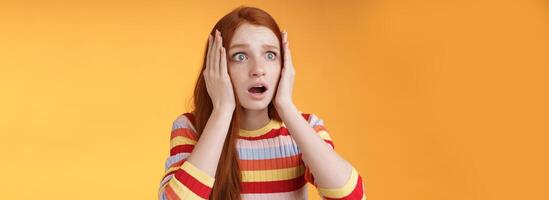 Worry insecure redhead european panicking girl grab head hands both sides gasping open mouth shocked stare frightened upset watching terrible accident standing concerned orange background photo