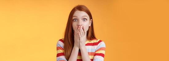 Shocked speechless impressed sensitive redhead european girl reacting stunning rumor gossiping find out secret gasping cover mouth palm stare camera astonished surprised, orange background photo