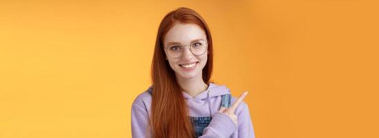 Friendly good-looking modern redhead young girl pointing left index finger showing awesome place suggesting go hang out smiling joyfully casually talking discuss new product, orange background photo