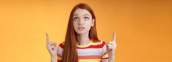 Stunned thrilled young redhead woman peer focused pointing up index fingers upwards look concentrated excited hold breath amused performance standing orange background intrigued and curious photo