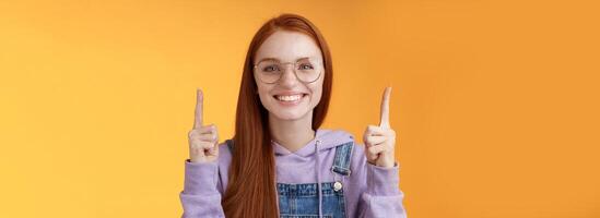 Lively charismatic pleasant redhead female student helpful pointing up index fingers showing awesome promo offer smiling delighted white teeth indicating advertisement recommend use product photo