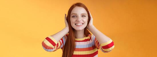 Cute playful tender smiling ginger girl blue eyes silly cover ears palms grinning fool around not hear anyone carefree unwilling listen noisy people standing careless relaxed orange background photo