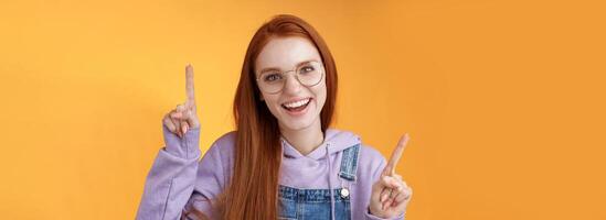 Lively enthusiastic millennial redhead girl coworker having fun celebrating small break joyfully dancing pointing up index fingers singing smiling white teeth promoting product, orange background photo