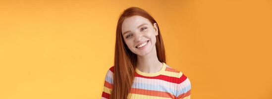 Pleasant charismatic young confident carefree redhead charming girl tilting head smiling broadly white teeth talking casually good mood express positive happy emotions standing orange background photo