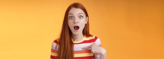 Shocked silly surprised redhead cute girl gasping drop jaw stunned stare camera full disbelief pointing herself cannot believe chosen picked participate event standing impressed orange background photo