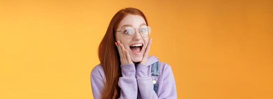 Excited happy smiling broadly redhead girlfriend drop jaw grinning thrilled look left astonished touch cheeks reacting awesome stunning news screaming happiness, standing orange background photo