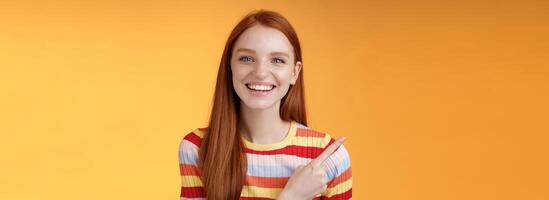 Happy charismatic smiling young ginger girl blue eyes sincere tender grin laughing broadly having fun discuss hilarious concert pointing upper left corner showing awesome product, orange background photo