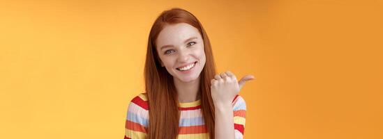 Friendly tender feminine kind redhead woman blue eyes pointing thumb left showing location where find product copy space gladly help discuss interesting promo grinning flirty, orange background photo