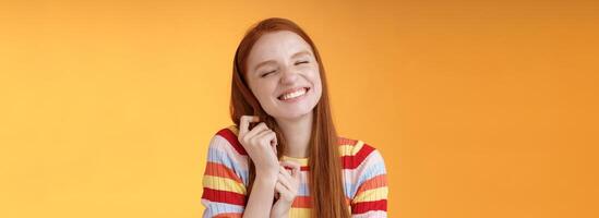 Happy dreamy romantic young tender ginger girl fantasizing creating love story imagination smiling broadly delighted close eyes touching hair strands recalling nice memory, standing orange background photo