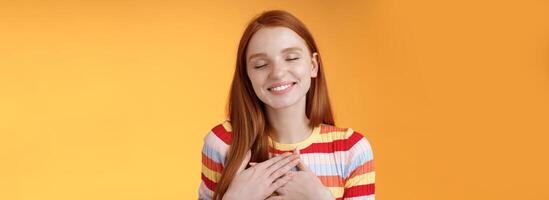 Happy romantic delighted sighing young lovely redhead girlfriend smiling broadly touching heart chest palms close eyes recalling date passionate kiss feel gratitude warmth, orange background photo