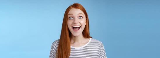 Surprised pleased happy impressed redhead european girl 20s reacting amused wide eyes look admiration joy receive incredible offer standing excited express thrill upbeat feelings, blue background photo