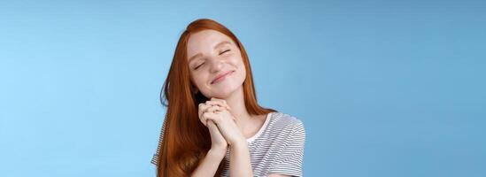 Charming delighted happy dreamy redhead girlfriend ginger long hair tilting head close eyes smiling pleased daydreaming thinking about sweet tender memories press palms together, blue background photo