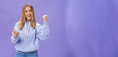 Yes we did it. Pleased excited woman in hoodie raising fist in celebration and success gesture smiling broadly triumphing holding smartphone reacting to positive news in cellphone over purple wall photo