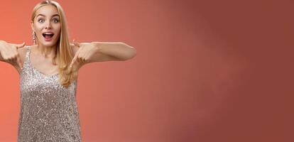 Impressed excited attractive glamour blond girl in silver glittering dress gasping thrilled pointing down glance camera fascinated check out fabulous awesome promo, standing amused red background photo