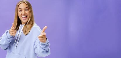 Enthusiastic and charismatic woman feeling joyful meeting friends checking out cool copy space pointing with finger guns at camera smiling broadly posing in stylish hoodie over purple wall photo