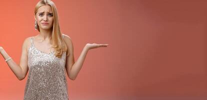 Nervous unsure doubtful cute blond woman struggle make decision shrugging pointing sideways frowning upset standing insecure feel pressure cannot decide choice make, frustrated red background photo