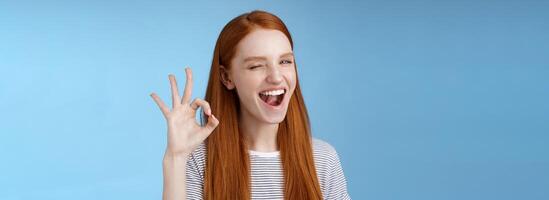 Sassy attractive redhead girl winking mysteriously smiling broadly give approval sign show okay ok excellent gesture satisfied good choice agree great decision, standing delighted blue background photo