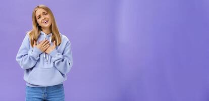 Sighing touched and pleased beautiful young girl with blond hair in hoodie holding palms on heart and smiling at camera thankful and grateful looking at heartwarming scene against purple wall photo