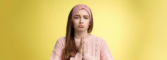 Clingy sad whining girlfriend bagging buy her dress grimacing pouting and frowning upset, pressing palms together in pray, asking favour, apologizing, praising friend to help over yellow background photo