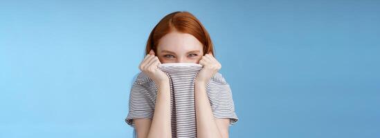 Silly flirty amused attractive playful redhead girlfriend hiding face pulling t-shirt head squinting devious mysteriously giggle laughing hope disguise pranking friend standing blue background photo