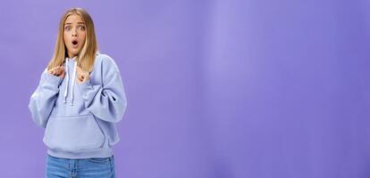 Woman scared of sudden loud noise in dark gasping looking shocked and concerned tilting backwards with frightened look pressing clenched fists to body from fear, wearing warm hoodie over purple wall photo