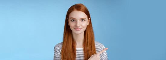 Attractive modest good-looking redhead girlfriend acting mature friendly smiling pointing left index finger giving direction show way bathroom standing blue background joyful kind grin photo