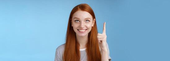 Intrigued lively happy smiling redhead european girl 20s look pointing up index fingers amused check out interesting sale holiday promo offer standing intrigued thrilled blue background photo