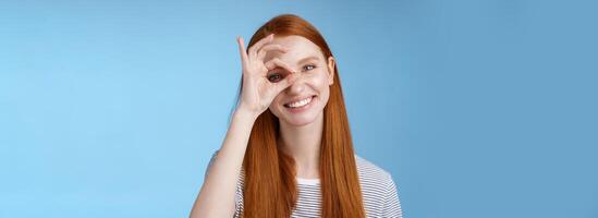 Charismatic happy adorable redhead teenage girl sincere eyes making circle eye show okay ok sign delighted like approve cool idea smiling satisfied achieve perfect score, standing blue background photo
