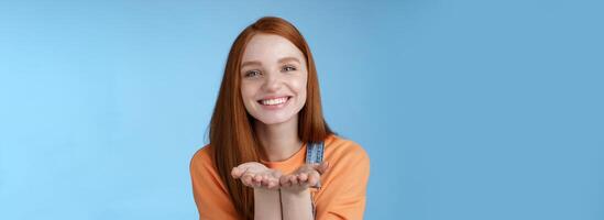 Cute tender kind young ginger girl giving all love you, hold something palms showing camera smiling delighted introduce present grinning romantic gesture send air kisses, blue background photo