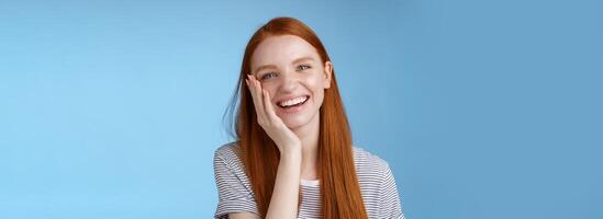 Charismatic talkative friendly-looking happy laughing redhead girl having fun discuss previous summer holidays make jokes chuckling touching face amused standing cheerful blue background photo