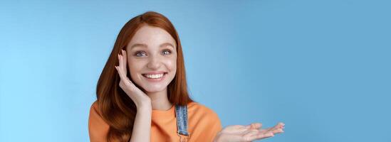 Cute tender chatismatic glad smiling redhead girl presenting awesome product show object palm hold hand raised blank copy space grinning impressed receive silly charming gift, blue background photo