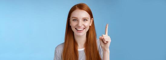 Got excellent idea pick for you. Attractive happy friendly-looking helpful redhead female shop assistant showing gorgeous dress customer pointing up index finger smiling glad help, blue background photo