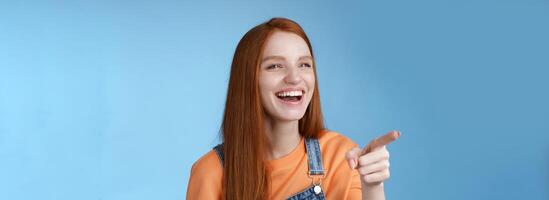 Adorable fascinated young attractive tender redhead amused girl having fun pointing looking left astonished laughing having fun see funny advertisement chuckling joyfully, blue background photo
