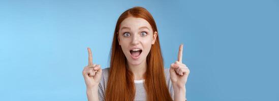 Surprised happy enthusiastic young redhead woman reacting impressed upper hanging promo pointing up index fingers drop jaw amused look thrilled camera telling about awesome offer, blue background photo