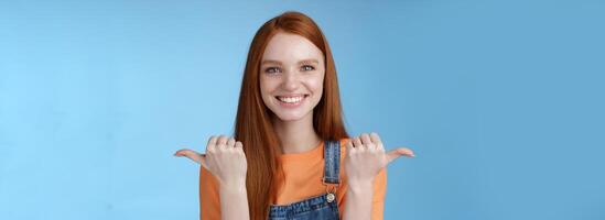 Indoor shot charismatic assertive happy smiling redhead woman orange shirt denim overalls pointing sideways thumbs left right showing choices opportunities give chance choose, blue background photo