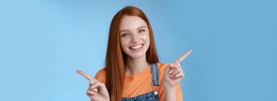 Friendly happy laughing pretty redhead girl female student showing lots opportunities advice make choice pointing sideways left right introducing different product grinning gladly, blue background photo