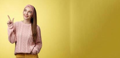 Portrait of amused interested cute 20s european girl wearing sweater, headband looking upper left corner pointing sideways smiling intrigued, ecstatic, captured by curious promo over yellow wall photo