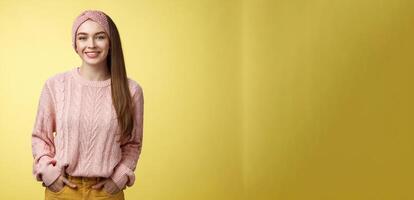 Charming friendly young girl in knitted sweater and headband holding hands in pockets, smiling, looking positive motivated achieving goals having great day, posing positive against yellow background photo