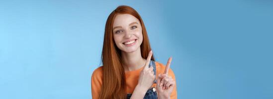 Happy tender redhead girl sincerely smiling white teeth helpful look camera excited give hand pointing upper left corner introduce sale offer recommend try promo standing blue background photo