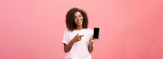 Suggest this model of phone. Happy charming african american curly-haired woman in stylish outfit holding smarpthone and pointing at device screen while recommending cool app over pink wall photo