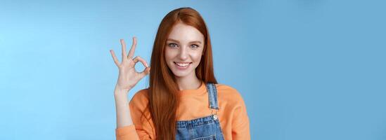 Sassy confident redhead girl got under control show okay ok excellent gesture cheeky look camera grinning assertive assuring everything awesome give permission approve good product, blue background photo