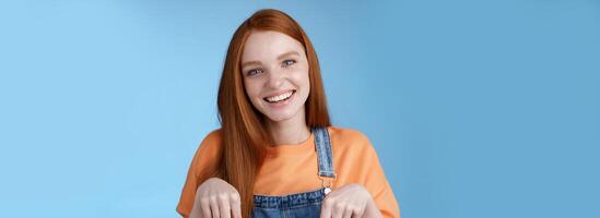 Friendly happy redhead girl smiling lively pretty grin pointing down index fingers offering good offer recommend use service standing blue background discuss interesting product, blue background photo