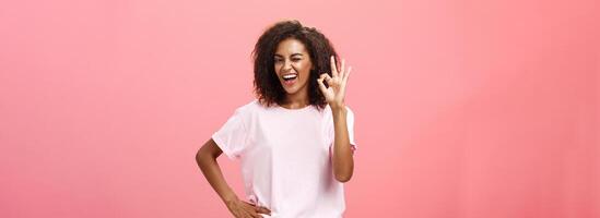 Girl got us covered. Charming charismatic and confident african american skillful woman in stylish outfit holding hand on waist showing okay gesture and winking assured at camera over pink wall photo