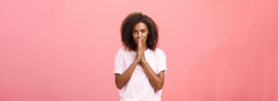 Creative and smart stylish attractive african american woman with curly hairstyle having great plan or idea feeling ambitious holding hands in pray over mouth gazing thoughtful at camera photo