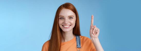 Confident happy charismatic ginger girl blue eyes freckles helpful showing product raising index finger up pointing cool copy space smiling broadly made choice recommend pick, studio background photo