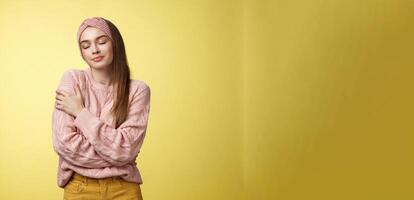 Sweet glamour young girl wearing warm comfy sweater embracing herself crossing arms over body in hug, smiling soft and kind close eyes, recalling lovely days, tender memories over yellow wall photo